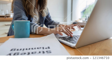 Young Freelance Woman Working from Home in Dining Room with Laptop and Coffee Mug, Focused on Strategic Planning Document 121346710