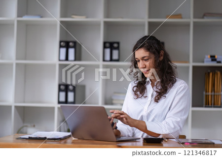 Confident Businesswoman Working on Laptop in Modern Office Environment with Shelves in Background 121346817