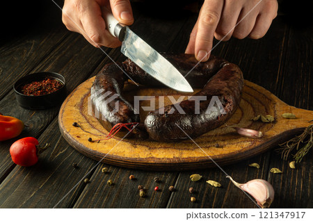A chef cuts blood sausage with a knife on a wooden breakfast cutting board. Close-up of the cook hands cutting the sausage surrounded by aromatic spices A chef cuts blood sausage with a knife on a wooden breakfast cutting board. Close-up of the cook hands cutting the sausage surrounded by aromatic spices 121347917