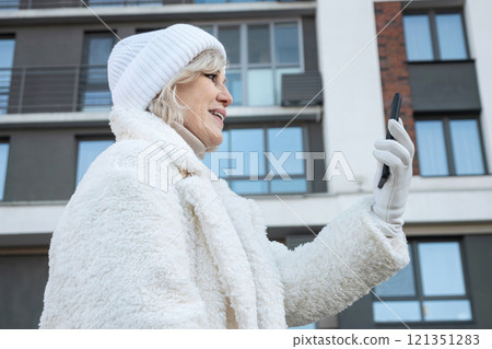 Smiling senior woman using smartphone wearing white winter clothes in city Smiling senior woman using smartphone wearing white winter clothes in city 121351283