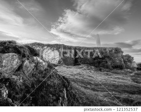 High-angle view of a coastal landscape. A slender white tower perches atop dark, stratified rocks. The sky are visible. High-angle view of a coastal landscape. A slender white tower perches atop dark, stratified rocks. The sky are visible. 121352537