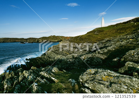 A white navigational marker is seen on a steep, rocky clifftop. The ocean is visible in the background, with a distant landmass. A white navigational marker is seen on a steep, rocky clifftop. The ocean is visible in the background, with a distant landmass. 121352539