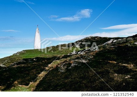 The image shows a white conical structure on a hill. The hill is a mix of grass and dark, rocky patches. The sky is mostly clear and blue. The image shows a white conical structure on a hill. The hill is a mix of grass and dark, rocky patches. The sky is mostly clear and blue. 121352540