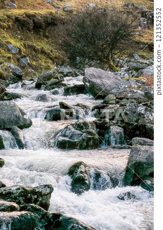 Clear water flows rapidly over a bed of dark rocks. A leafless tree sits among rocks beside the stream. The land slopes gently upward to distant hills. Clear water flows rapidly over a bed of dark rocks. A leafless tree sits among rocks beside the stream. The land slopes gently upward to distant hills. 121352552