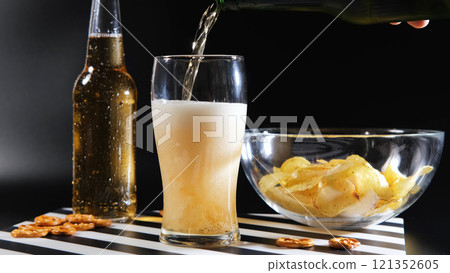 Frosty beer poured into a glass with chips and pretzels on a dark background 121352605