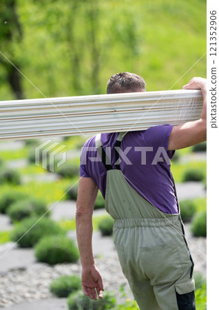 A male worker carries boards for sheathing a gazebo in a lavender field. A male worker carries boards for sheathing a gazebo in a lavender field. 121352906