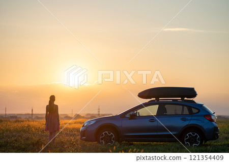 Dark silhouette of lonely woman relaxing near her car on grassy meadow enjoying view of colorful sunrise. Young female driver resting during road trip beside SUV vehicle. 121354409