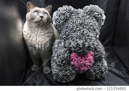 A cute gray cat sitting next to a decorative teddy bear made of gray roses with a pink heart. The scene is set on a black background, ideal for themes like pets, decor, or Valentine's Day. 121354802