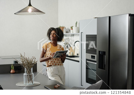 African American woman with tablet in hands looking at kitchen lamp while setting bulb brightness 121354848