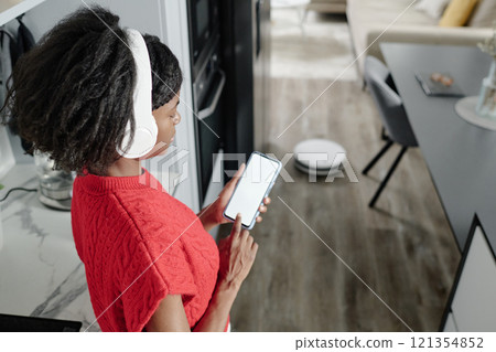 High angle shot of African American woman listening to music while controlling robot vacuum cleaner via smartphone app High angle shot of African American woman listening to music while controlling robot vacuum cleaner via smartphone app 121354852