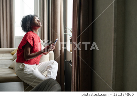 African American woman sitting on couch near huge window while using smartphone in her free time 121354860