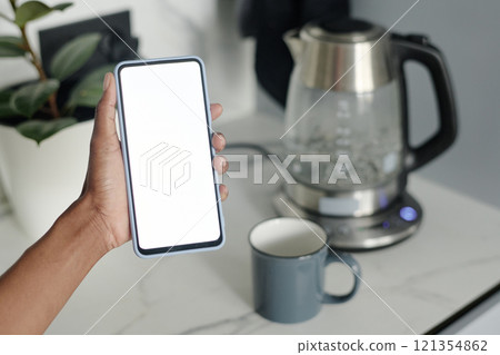 High angle hot of smartphone with white screen in unrecognizable female hand, boiling electric glass kettle in background High angle hot of smartphone with white screen in unrecognizable female hand, boiling electric glass kettle in background 121354862