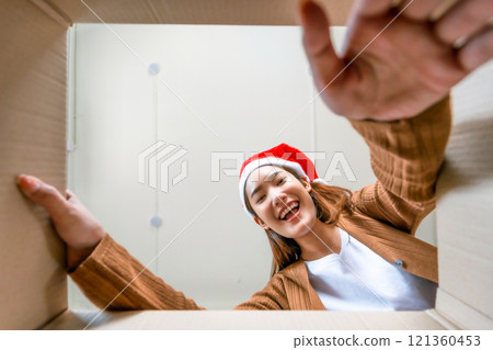 Low angle view of surprised young Asian woman wearing Christmas hat and unpacking 121360453