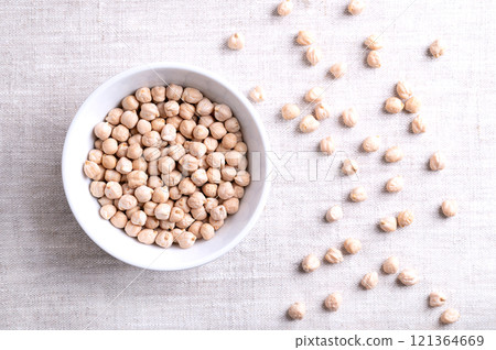 Dried chickpeas in a white bowl, on linen fabric, from above. Whole and light tanned chick peas, a high in protein legume and fruit of Cicer arietinum. Key ingredient in hummus and in chana masala. 121364669