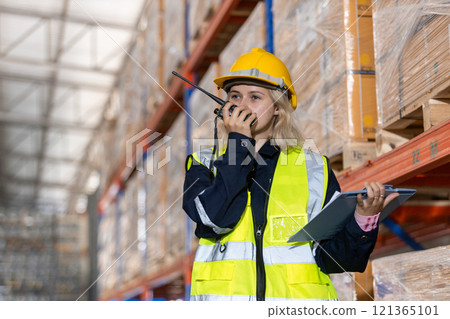Female blonde hair professional worker wearing safety uniform and hard hat holding clipboard and walkie-talkie inspect goods on shelves in warehouse. Worker check stock inspecting product in factory. 121365101