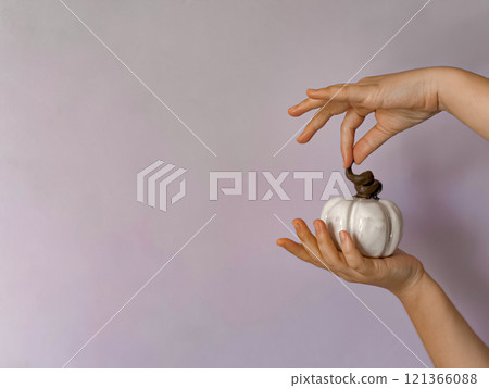 Close up of hand holding small white ceramic pumpkin with twisted brown stem against soft lavender background with copy space. Minimalist and symbolic representation of autumn, Halloween fall decor. Close up of hand holding small white ceramic pumpkin with twisted brown stem against soft lavender background with copy space. Minimalist and symbolic representation of autumn, Halloween fall decor. 121366088