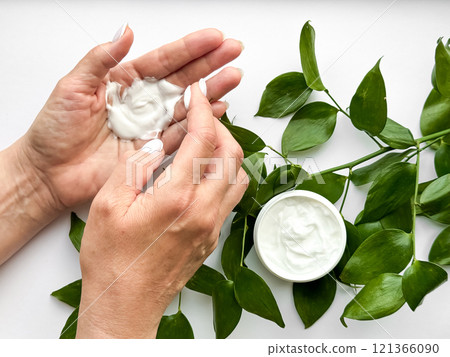 Close up of middle age woman hands applying hand cream with green leaves on white background. Skincare and natural beauty concept. For healthcare, wellness, and organic product poster. Close up of middle age woman hands applying hand cream with green leaves on white background. Skincare and natural beauty concept. For healthcare, wellness, and organic product poster. 121366090