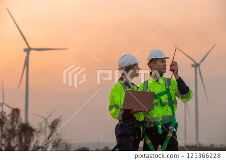 Team engineer wearing safety uniform holding laptop discussed plan about renewable energy at station energy power wind in evening when sunset. Technology environment reduce global warming problems. 121366228