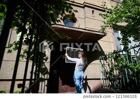 Woman holding photovoltaic solar panel in front of historical building, wearing plaid shirt and sunglasses. Concept of integration of sustainable renewable energy sources into old architecture. 121366328