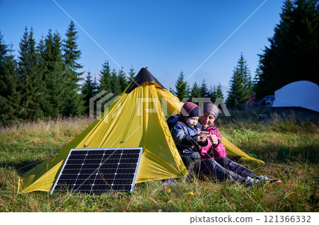 Young child and mother play with smartphone while charging with photovoltaic solar panel near tourist tent in summer. Integration of renewable energy in outdoor camping activities. 121366332