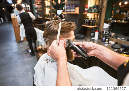 Professional barber using comb and electric shaver to cut side hair. Close-up view of male customer getting stylish haircut. Side view of man on background of other hairdresser. 121366357