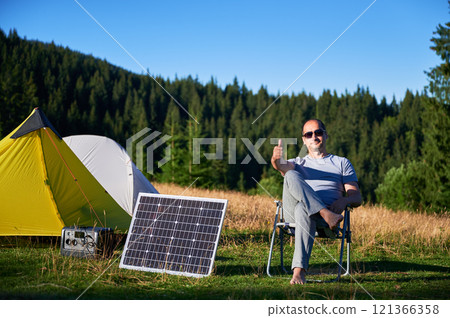 Man using photovoltaic solar panel for charging portable power station outdoor in eco-friendly camping. Male tourist giving thumbs up near tents. On background tall pine trees and clear blue sky. 121366358