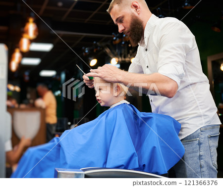 Man barber cutting little boy's hair using comb and scissors. Child getting haircut from adult male, likely barber. Professional hairdresser and cute client at modern barbershop. 121366376