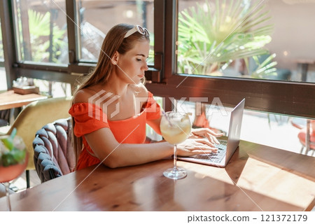 Woman works on laptop in a sunny cafe, glass of juice nearby. The setting is modern, with natural light illuminating the space. The image captures a moment of focused work. 121372179