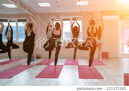 A group of six athletic women doing pilates or yoga on pink mats in front of a window in a beige loft studio interior. Teamwork, good mood and healthy lifestyle concept. A group of six athletic women doing pilates or yoga on pink mats in front of a window in a beige loft studio interior. Teamwork, good mood and healthy lifestyle concept. 121372183