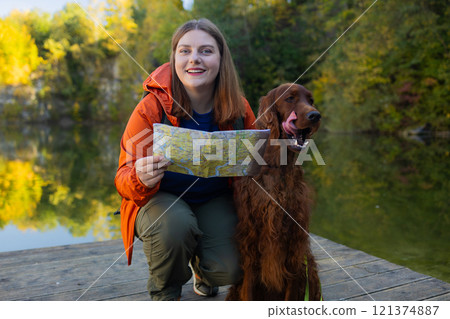Young woman traveling with dog. Beautiful female sitting near a lake together with Irish Setter dog and looking around. Happy girl interact with her pet at hiking rest with mountain and forest view. Young woman traveling with dog. Beautiful female sitting near a lake together with Irish Setter dog and looking around. Happy girl interact with her pet at hiking rest with mountain and forest view. 121374887