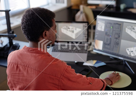Person wearing red shirt designing architectural floor plans on computer screens in modern office environment. Open window with natural lighting entering room Person wearing red shirt designing architectural floor plans on computer screens in modern office environment. Open window with natural lighting entering room 121375598
