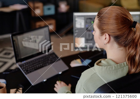 Person sitting at desk and observing design layout on computer screen, focusing attentively. Workspace includes devices like laptop and desktop, contributing to a professional environment Person sitting at desk and observing design layout on computer screen, focusing attentively. Workspace includes devices like laptop and desktop, contributing to a professional environment 121375600