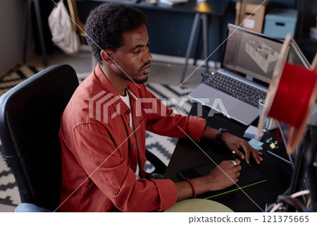 Man sitting at a desk with CAD software on screen, creating intricate 3D models for tech project. Surroundings include a modern office space with various tools and gadgets Man sitting at a desk with CAD software on screen, creating intricate 3D models for tech project. Surroundings include a modern office space with various tools and gadgets 121375665