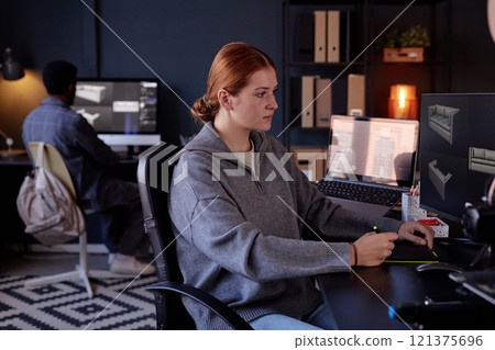 Portrait of woman sitting at desk in modern office working on computer with another person working in background, in the open workspace with multiple screens and shelves Portrait of woman sitting at desk in modern office working on computer with another person working in background, in the open workspace with multiple screens and shelves 121375696