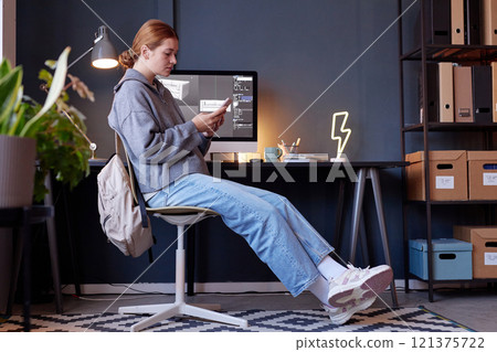 Young woman sitting at desk with mobile phone focused on screen, wearing casual clothes in cozy home office setup. Shelves with boxes, decor, and potted plant in background Young woman sitting at desk with mobile phone focused on screen, wearing casual clothes in cozy home office setup. Shelves with boxes, decor, and potted plant in background 121375722