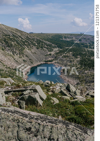 Stone hillsides, evergreen conifers, rocks in distance and emerald colored lake. Summer day in low mountains. 121375758