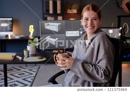Smiling woman holding mug while sitting in design office with computer screens displaying 3D models in background portraying creative work environment Smiling woman holding mug while sitting in design office with computer screens displaying 3D models in background portraying creative work environment 121375969
