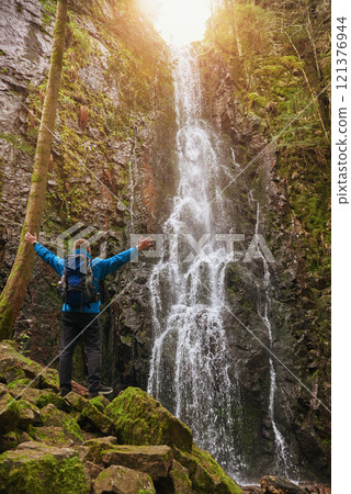 Tourist attraction of Germany - falls of Burgbach Waterfall near Schapbach, Black Forest, Baden-Wurttemberg, Germany. Man hiker in blue jacket standing on stone and looks at flow of falling water Tourist attraction of Germany - falls of Burgbach Waterfall near Schapbach, Black Forest, Baden-Wurttemberg, Germany. Man hiker in blue jacket standing on stone and looks at flow of falling water 121376944