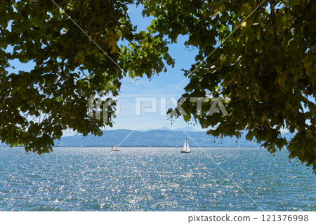 Picturesque View of Bodensee Through Tree Arch in Lindau Showcasing White Sailboats and Swiss Alps. Idyllic Summer Day Captures European Vacation Paradise with Sailing, Nature, and Mountain Vistas. 121376998