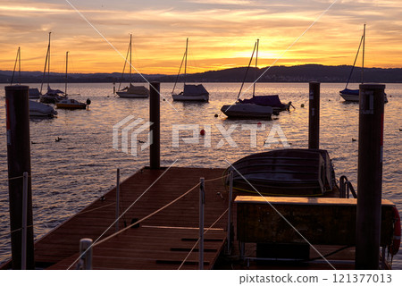 A serene sunset over a calm river, with vibrant orange and pink hues in the sky. A small sailboat is anchored in the water, reflecting the colorful sky. 121377013
