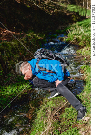 Hiker drinking stream water in mountain. Adult man drinking water from the mountain creek. Tourist wearing casual clothes and sunglasses making a sip of mountain river water from the palms of his Hiker drinking stream water in mountain. Adult man drinking water from the mountain creek. Tourist wearing casual clothes and sunglasses making a sip of mountain river water from the palms of his 121377021