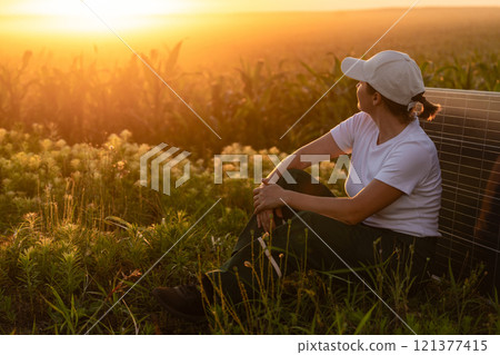 Woman wearing white cap sits next to solar panel at sunset. Wind turbines in the background. Woman wearing white cap sits next to solar panel at sunset. Wind turbines in the background. 121377415
