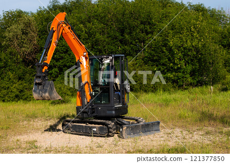 Excavator on a construction site next to a blooming field. Field with green grass and a small crawler excavator standing nearby 121377850