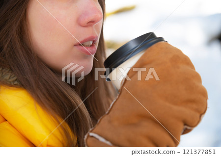 Cute girl on the street in winter smiling and holding a disposable cup of coffee, close-up of the cup and her lips in her hands wearing mittens Cute girl on the street in winter smiling and holding a disposable cup of coffee, close-up of the cup and her lips in her hands wearing mittens 121377851