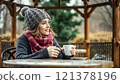 A girl in a knitted hat and scarf drinks hot tea in a gazebo 121378196
