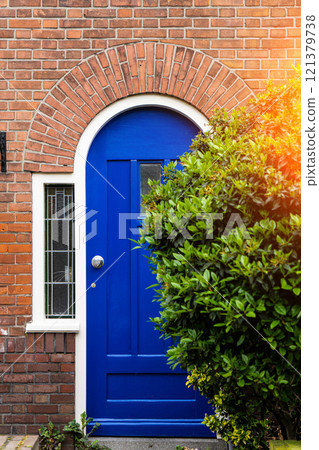 Wooden front door of a home. Front view of a wooden front door on a brick house with windows plants and a wide view of the porch and front walkway. Horizontal shot. 121379738