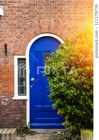 Wooden front door of a home. Front view of a wooden front door on a brick house with windows plants and a wide view of the porch and front walkway. Horizontal shot. 121379739