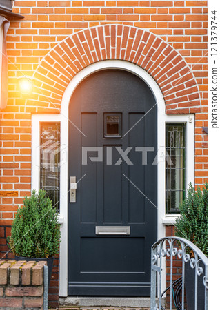 Wooden front door of a home. Front view of a wooden front door on a brick house with windows plants and a wide view of the porch and front walkway. Horizontal shot. 121379744
