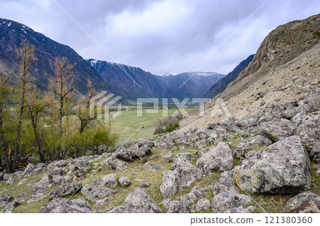 View from the top of a deep canyon surrounded by mountains, a river flowing below and a steep winding road descending down to the river on an overcast spring day. Chulyshman river valley, Altai, View from the top of a deep canyon surrounded by mountains, a river flowing below and a steep winding road descending down to the river on an overcast spring day. Chulyshman river valley, Altai, 121380360