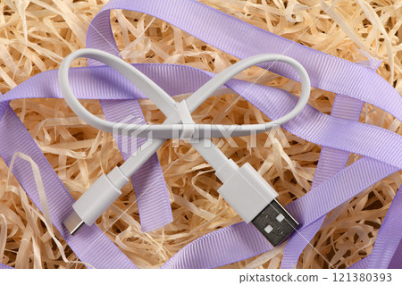 White USB cable in the form of a bow on the background of blue ribbon and decorative shavings. 121380393